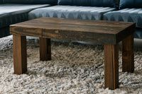 A handcrafted square coffee table, expertly built from sustainable reclaimed wood with a rich dark finish, anchors a cozy beige rug before a dark blue sofa—showcasing enduring quality in reclaimed wood furniture.