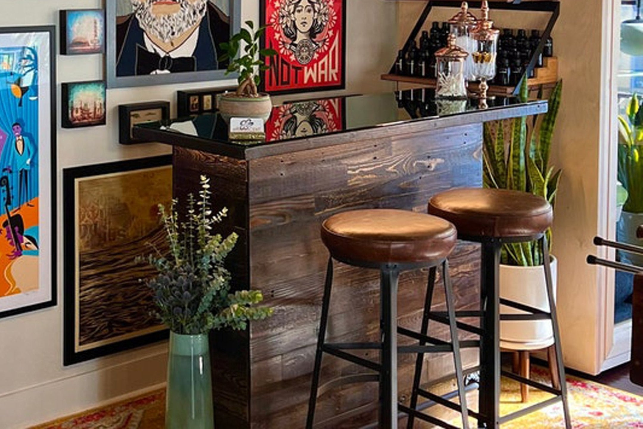 This handcrafted reclaimed wood bar table features a rustic counter and two brown leather stools. Surrounded by vibrant artwork, plants, and curated shelves, this Arizona-made piece brings enduring quality to any space.