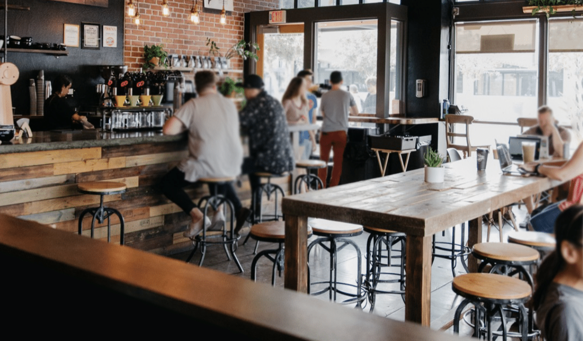 Bar-height reclaimed wood dining tables and stools in a cozy café setting, with natural light from large windows highlighting handcrafted Arizona furniture and sustainable materials; patrons seated at tables.