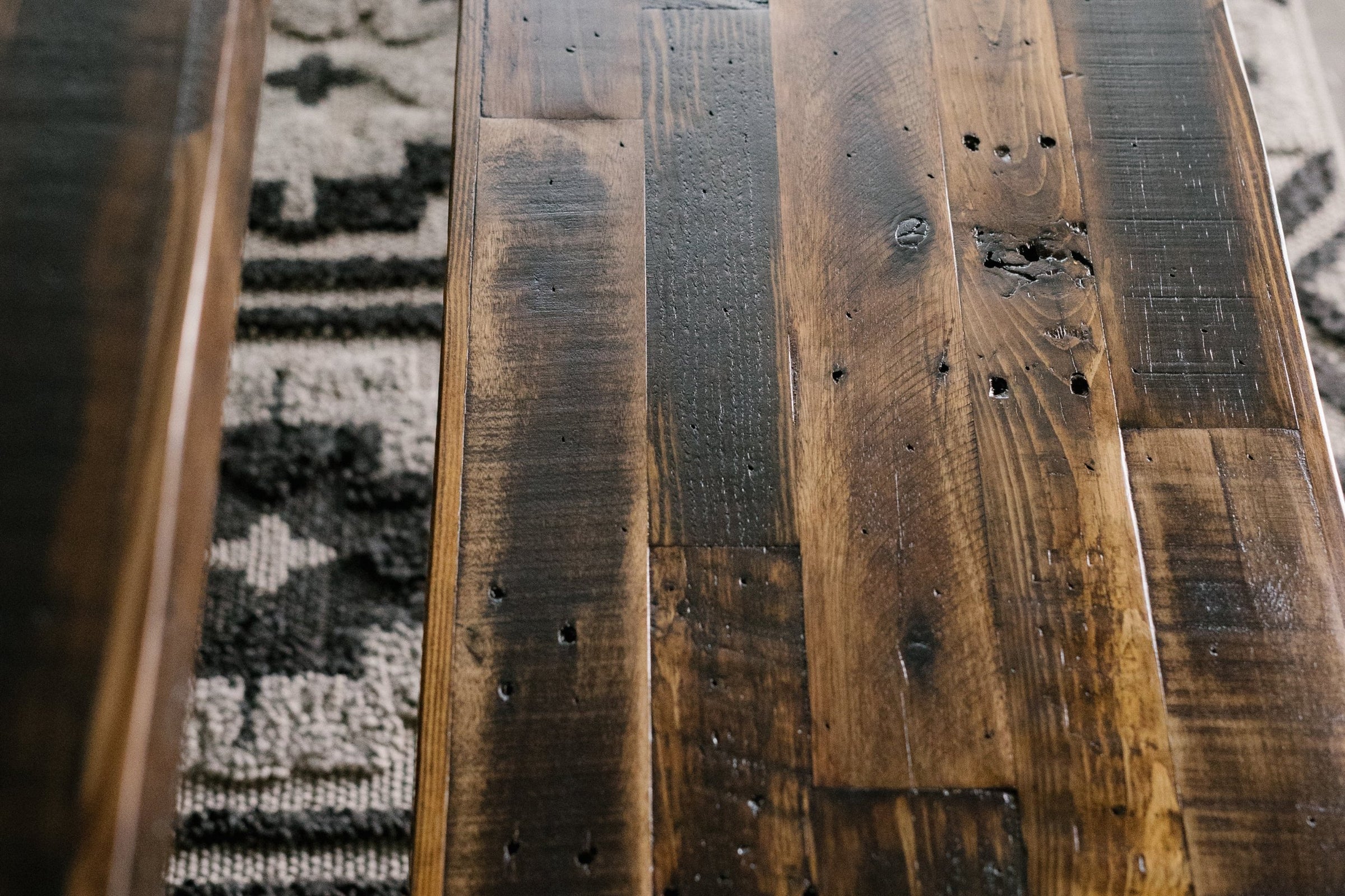 Close-up of a handcrafted reclaimed wood bench, showcasing weathered grain, natural knots, and artisan imperfections. A hint of patterned rug beneath emphasizes the enduring quality of this sustainable reclaimed wood furniture piece.