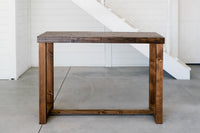 Experience timeless design with our handcrafted Sequoia counter-height table, made in Arizona from sustainably sourced reclaimed wood. Perfect as a narrow kitchen island or restaurant bar table, set on a concrete floor by a white wall and staircase.