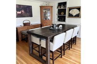 A welcoming dining space featuring a handcrafted Sequoia bar-height table, made from reclaimed wood with a rich, dark finish. Surrounded by six white upholstered chairs, it’s complemented by wooden floors and artisan cabinetry.
