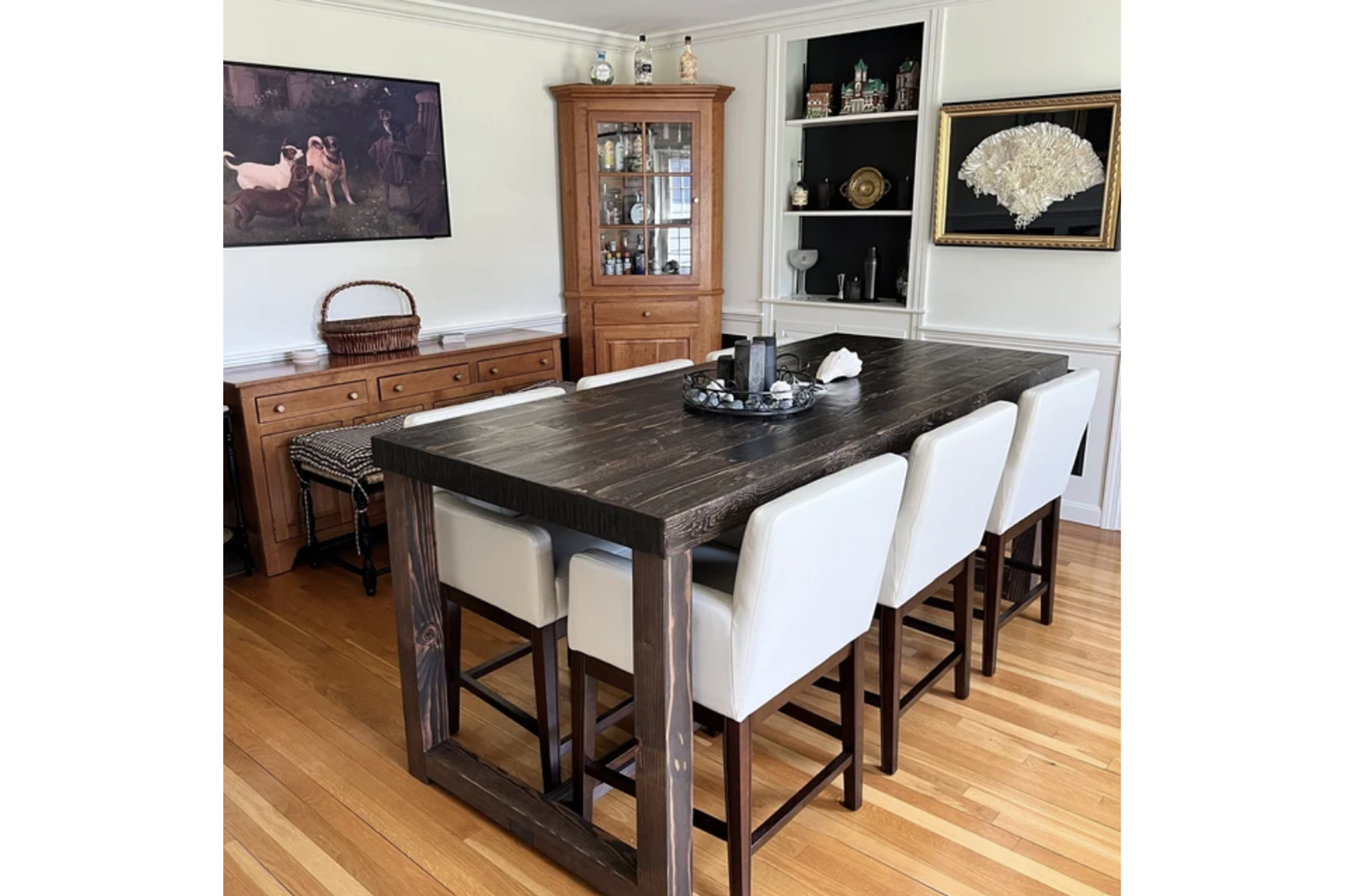 A welcoming dining space featuring a handcrafted Sequoia bar-height table, made from reclaimed wood with a rich, dark finish. Surrounded by six white upholstered chairs, it’s complemented by wooden floors and artisan cabinetry.