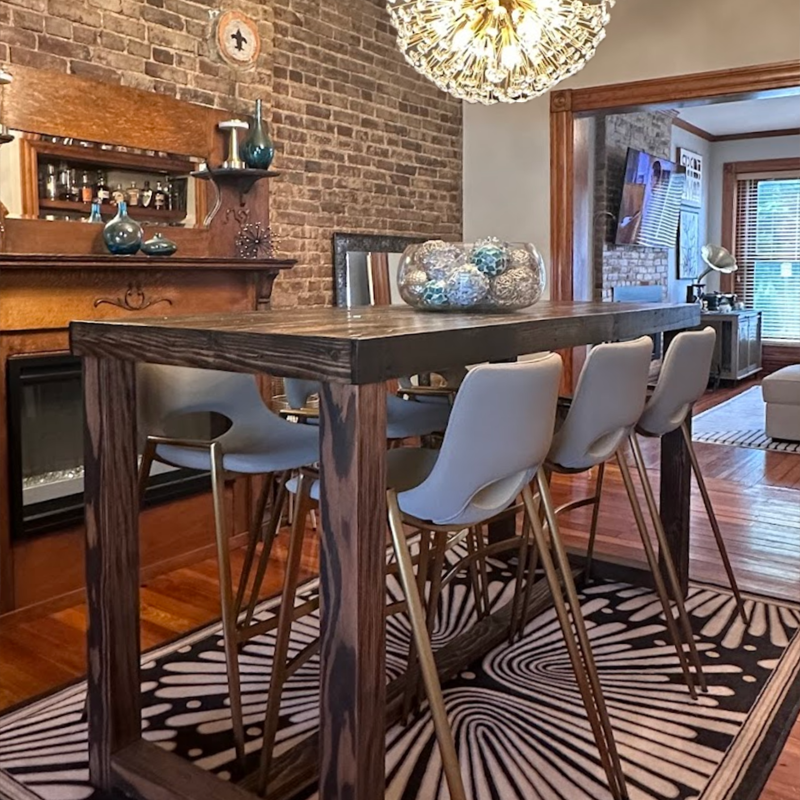 Bar-height dining table handcrafted in Arizona from reclaimed wood surrounded by six white chairs, set on a patterned rug with exposed brick walls and a modern chandelier in a contemporary dining room.