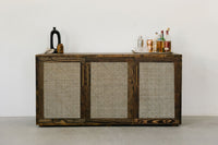 This handcrafted cane door sideboard, crafted in Arizona from sustainable materials, stands beside a white wall. Styled as a minimalist bar table with bottles and glassware, it exemplifies enduring quality and reclaimed wood furniture design.