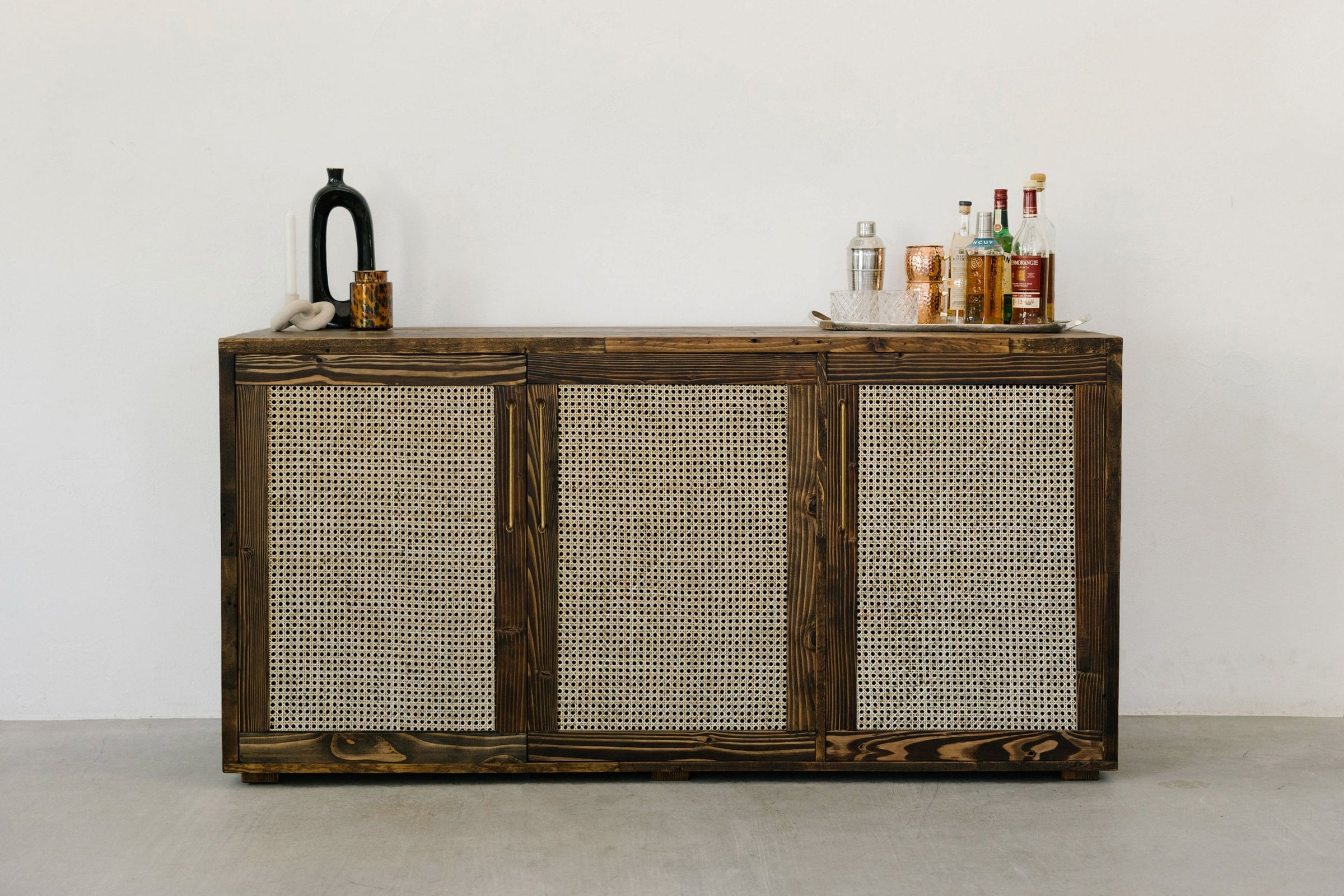 This handcrafted cane door sideboard, crafted in Arizona from sustainable materials, stands beside a white wall. Styled as a minimalist bar table with bottles and glassware, it exemplifies enduring quality and reclaimed wood furniture design.