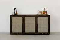 This handcrafted dark wood sideboard features woven rattan panels and is ideal as a reclaimed wood furniture piece or kitchen island. Accented with a black vase, small jar, and glassware for timeless, sustainable style.