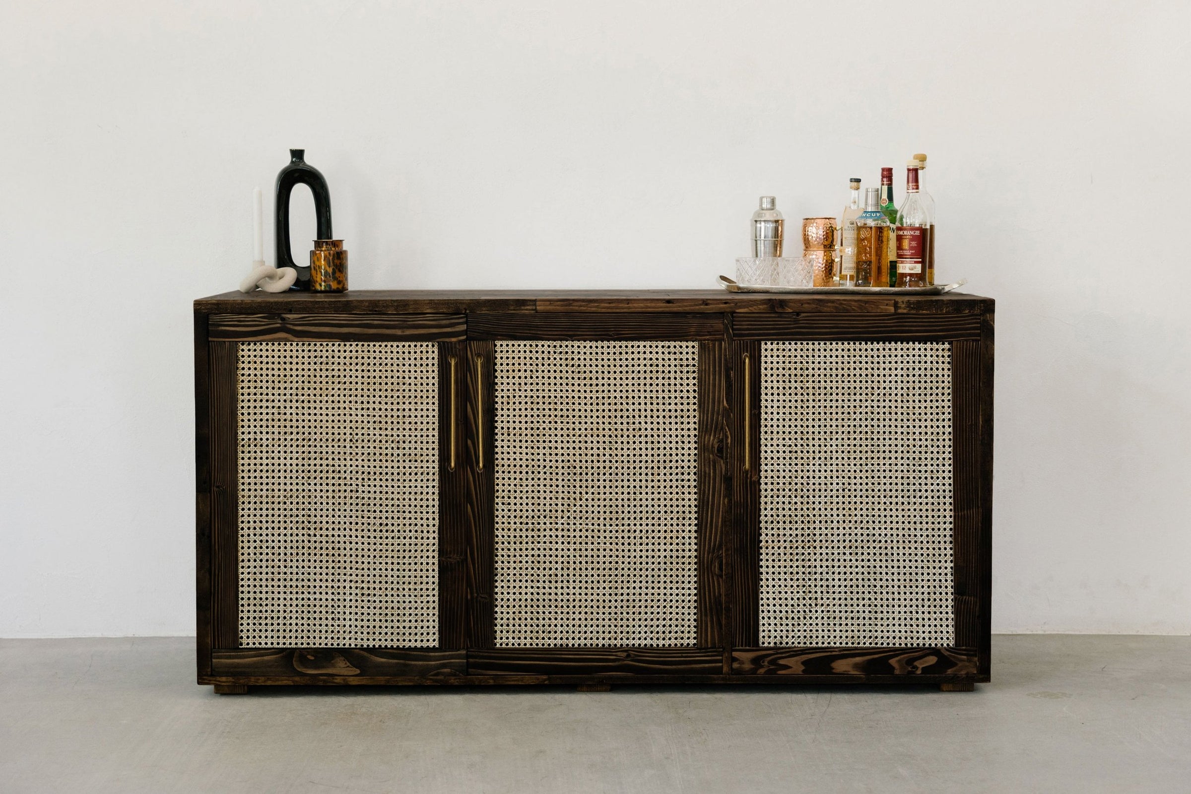 This handcrafted dark wood sideboard features woven rattan panels and is ideal as a reclaimed wood furniture piece or kitchen island. Accented with a black vase, small jar, and glassware for timeless, sustainable style.