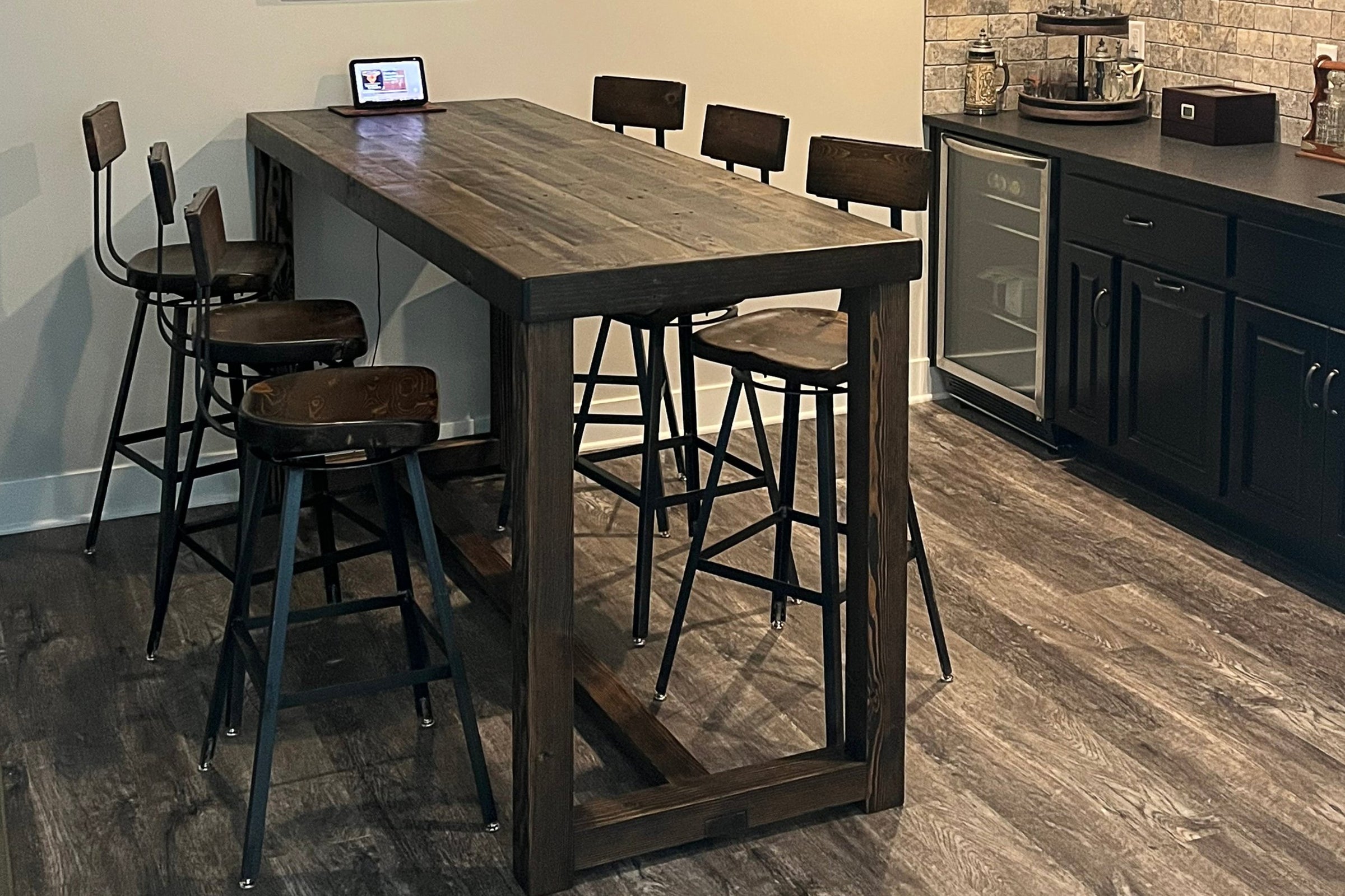 A modern kitchen showcases a custom Sequoia bar-height table, handcrafted in Arizona from reclaimed wood and steel. Six artisan stools encircle the table, with a tablet atop and coffee station set against rich dark wood flooring.