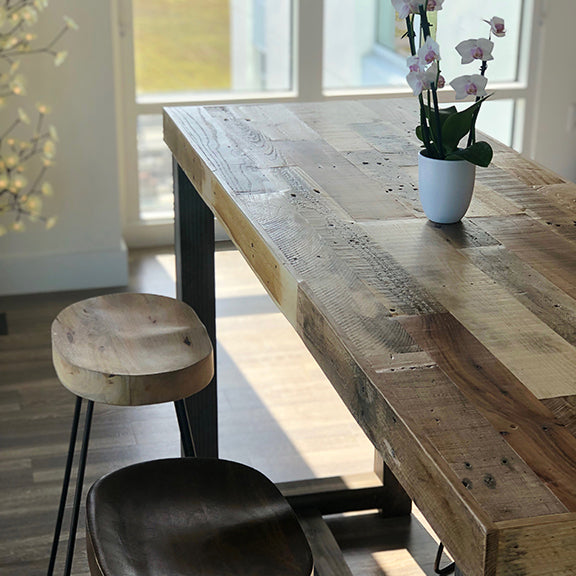 Bar-height dining table handcrafted in Arizona from reclaimed wood with steel legs, paired with two wooden stools; white potted orchid centerpiece by sunlit window—ideal for modern cafés or historic homes.
