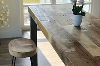 This handcrafted reclaimed wood bar-height table with a matching round stool embodies enduring quality and sustainable craftsmanship. Sunlight highlights the sequoia top, while a potted orchid adds a refined touch—perfect for kitchens or restaurants.