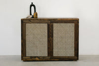 This handcrafted reclaimed wood sideboard features two rattan-panel doors, showcasing enduring quality and sustainable materials. Ideal as narrow bar table or kitchen island accent. Styled with a black vase, amber container, and décor piece.