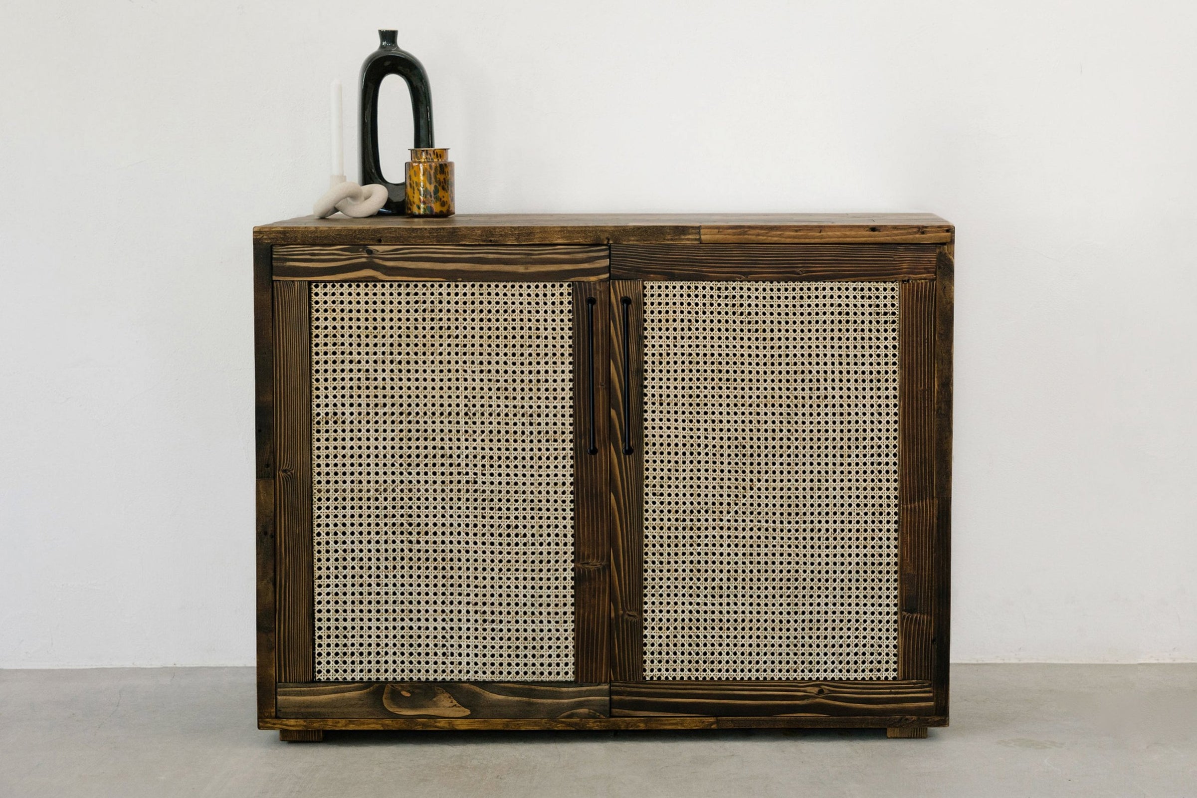 This handcrafted reclaimed wood sideboard features two rattan-panel doors, showcasing enduring quality and sustainable materials. Ideal as narrow bar table or kitchen island accent. Styled with a black vase, amber container, and décor piece.
