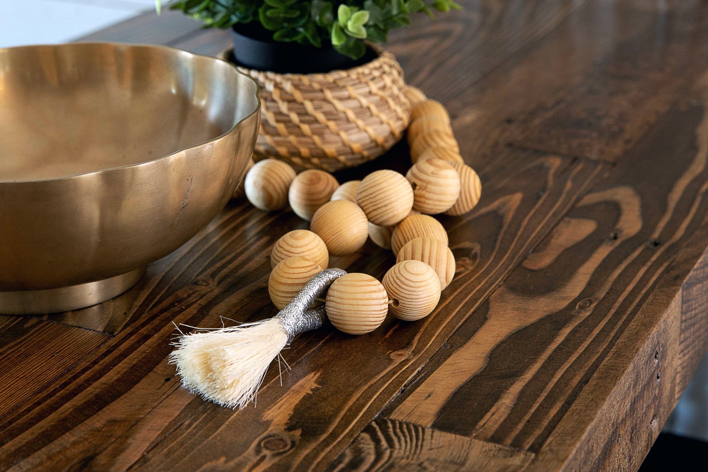 A handcrafted wooden beaded garland with a white tassel accents a reclaimed wood bar-height table, paired with a brass bowl and wicker plant pot. Showcasing enduring quality and sustainable materials for elevated spaces.