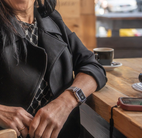 Handcrafted reclaimed wood cafe table with steel legs in a modern café setting, woman in black jacket seated with coffee and smartphone; Arizona-made sustainable furniture with enduring quality.