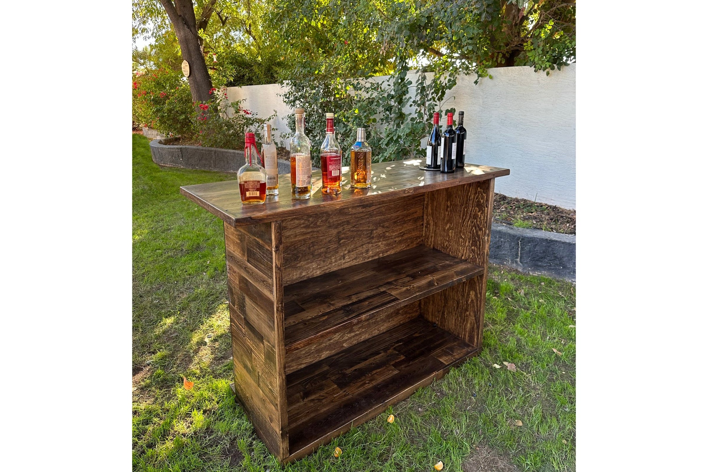 Crafted from reclaimed wood, this bar-height table showcases Arizona artisan skill and eco-friendly design. Perfect as a kitchen island or restaurant table, its durable surface displays curated bottles outdoors by a lush yard and white fence.