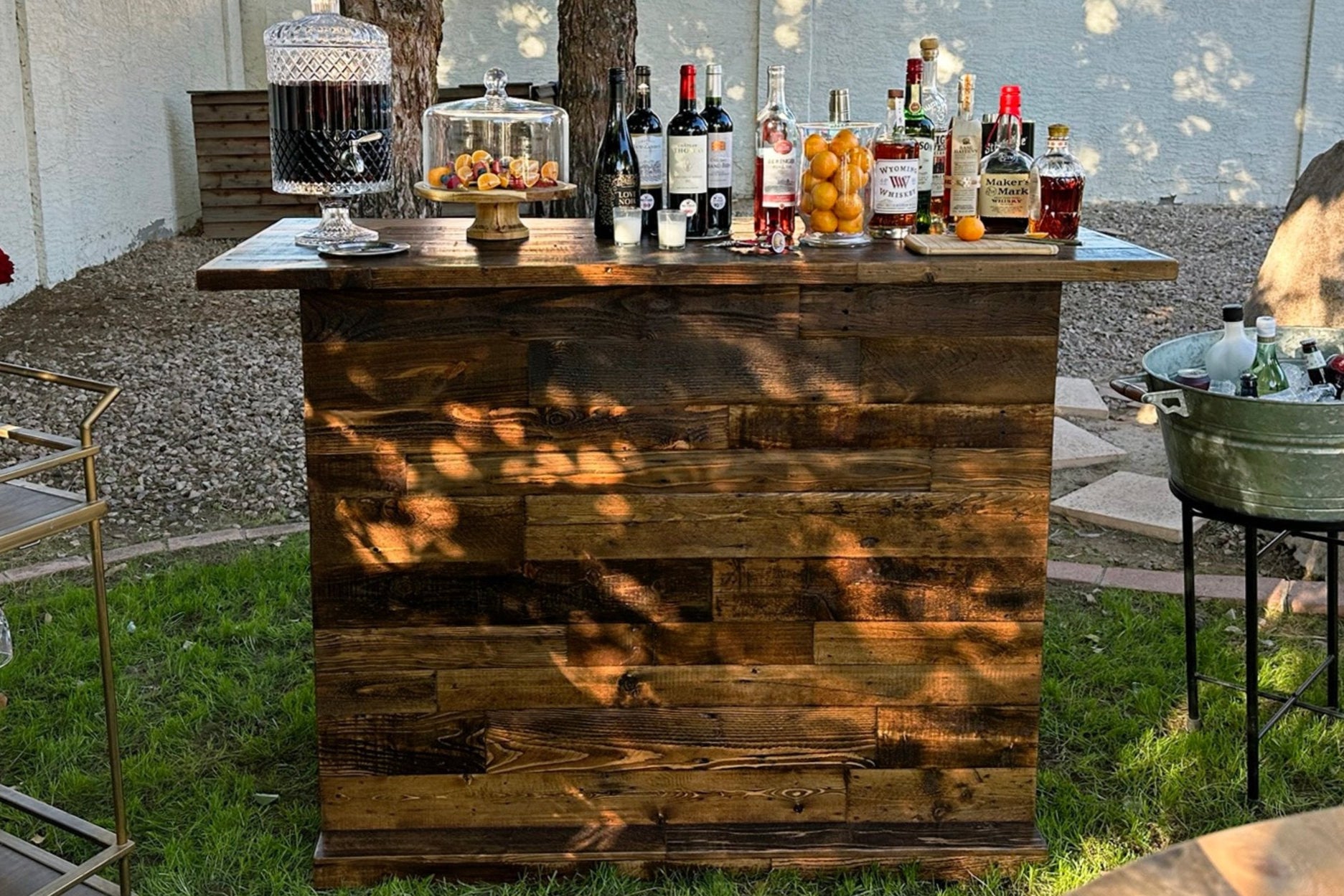 A handcrafted reclaimed wood bar-height table, expertly crafted in Arizona, stands on grass. Topped with liquor bottles, glassware, and snacks, this sustainable bar exudes warmth and enduring quality. Dappled sunlight enhances its charm.