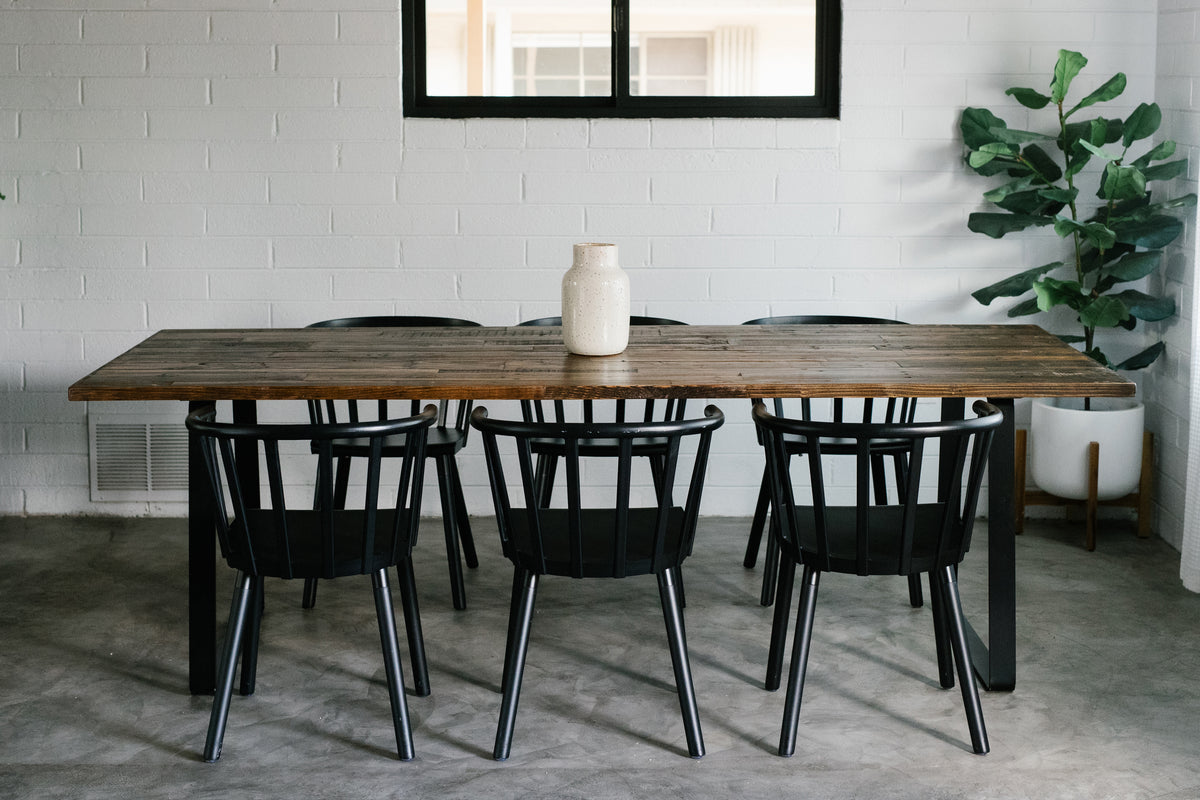 A minimalist dining space highlights a handcrafted Bell Dining Table in sustainably sourced reclaimed wood with a Provincial finish, paired with six black chairs. Ideal as a narrow dining table or restaurant table, accented by artisan details.