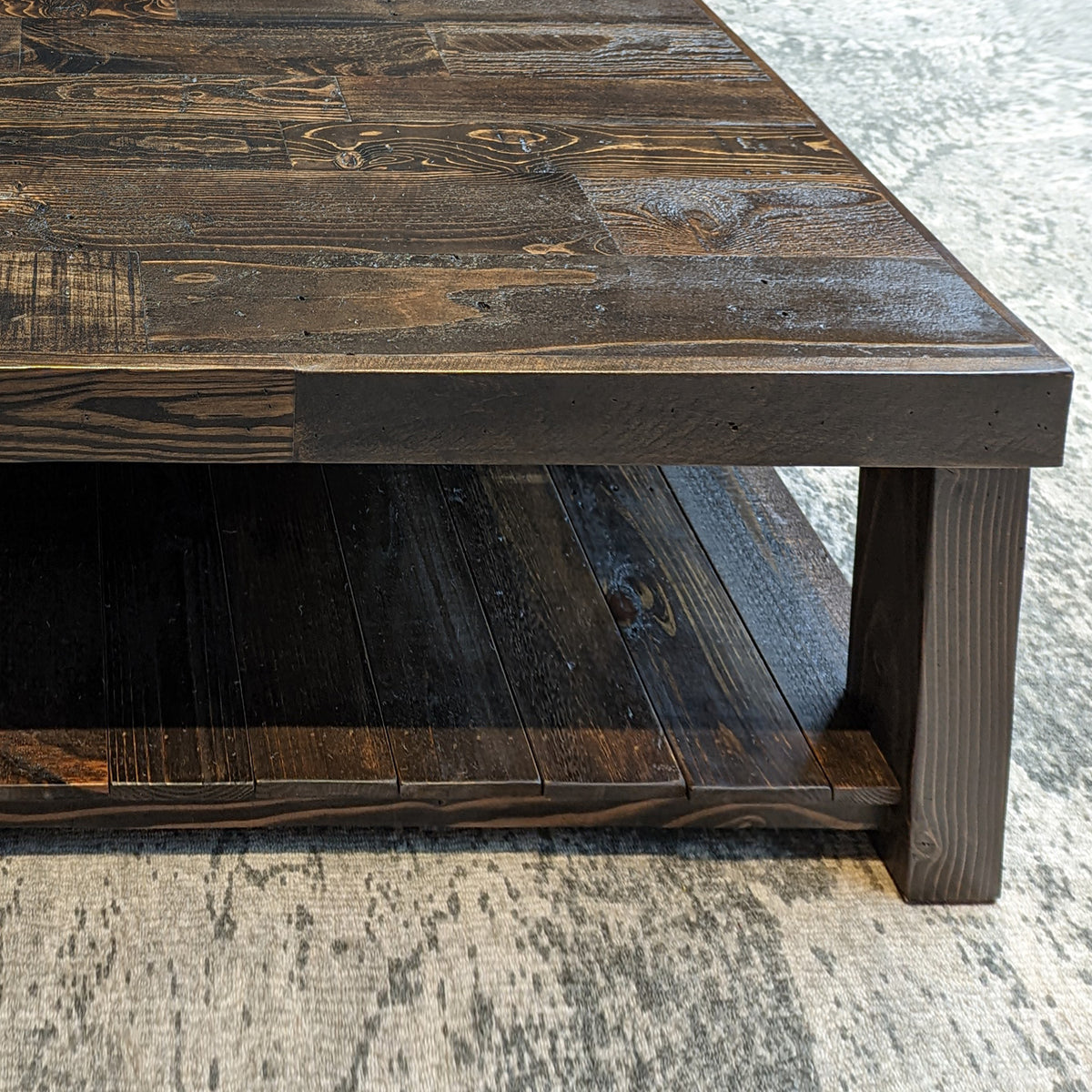 Close-up of a handcrafted dark reclaimed wood coffee table with lower shelf, shown on a light textured rug; ideal for modern living rooms, minimalist spaces, or historic homes.