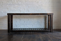 Expertly handcrafted in Arizona, this narrow bar-height console table features a slatted lower shelf and stands out against a white brick wall. Made from sustainable reclaimed wood, it’s ideal as a kitchen island or restaurant table.