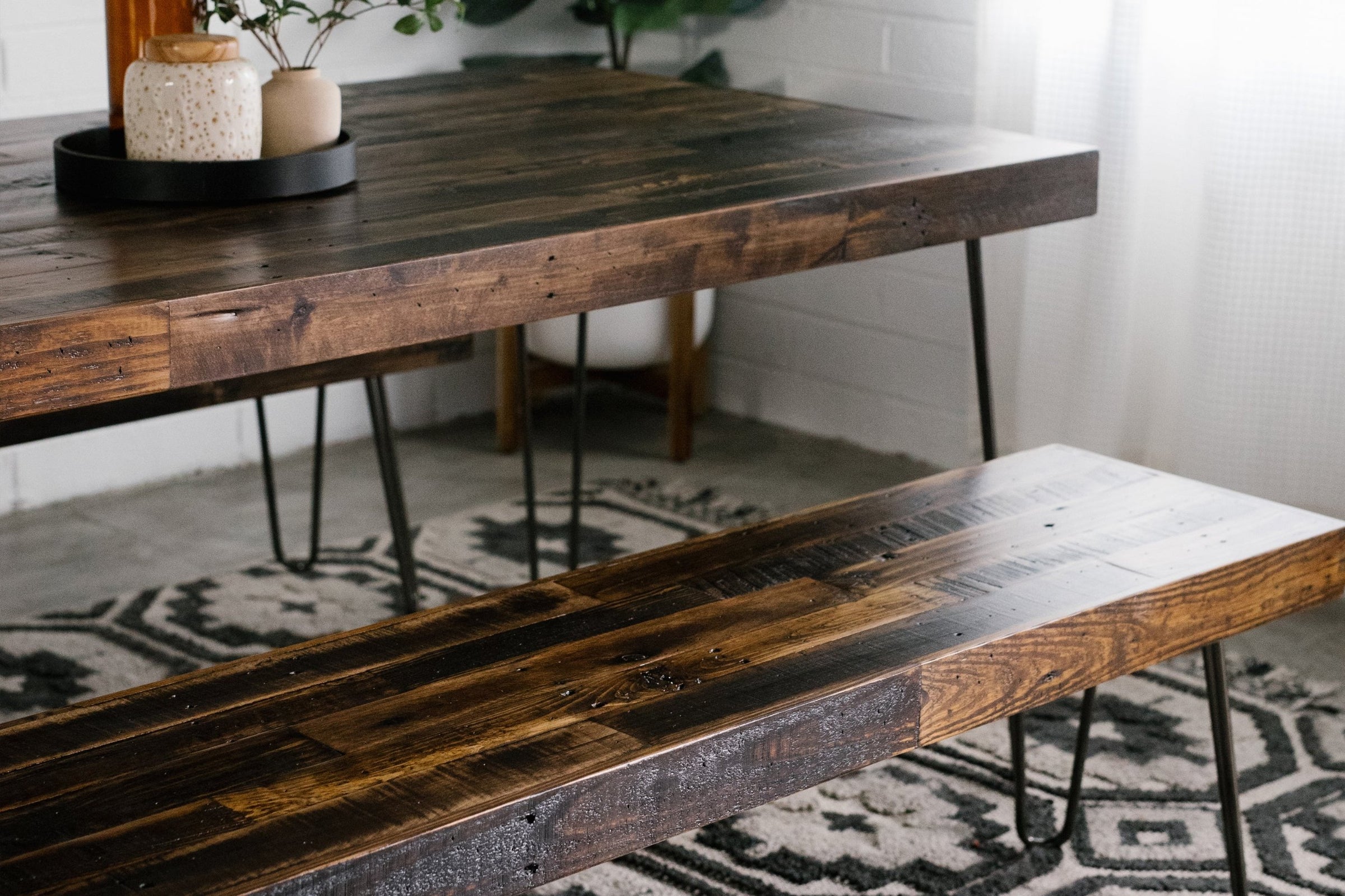 A close-up of a handcrafted dining table and bench with steel hairpin legs, made from sustainable reclaimed wood. Set on a patterned rug amid lush plants and sunlight, this Arizona-made table adds lasting quality to any home or restaurant.