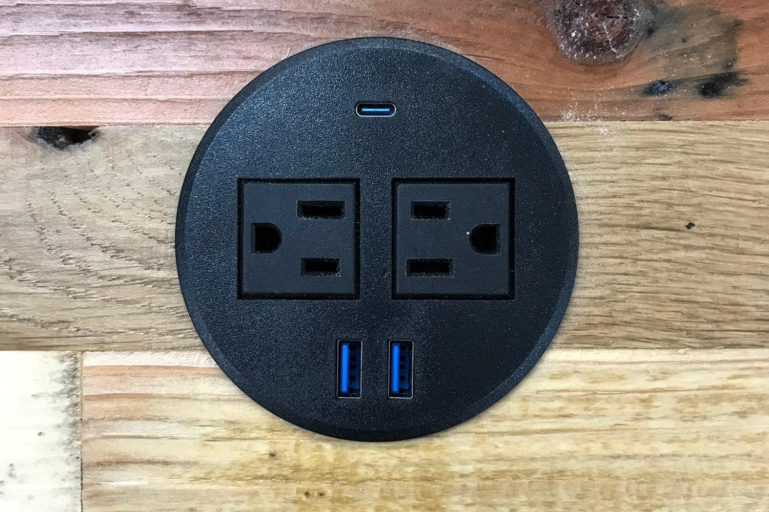 A round black power grommet set into a reclaimed wood surface, featuring two electrical sockets, blue USB ports, and a USB-C port—an ideal complement to handcrafted bar tables or sustainable kitchen islands made in Arizona.