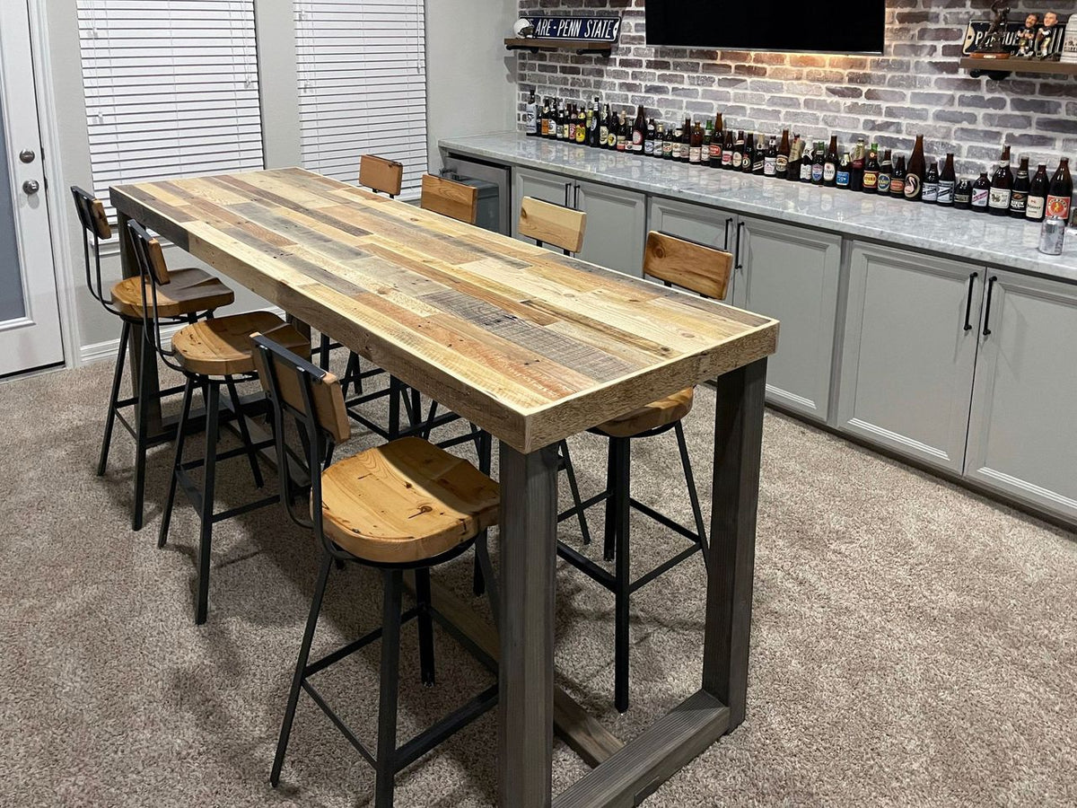 Bar-height dining table with reclaimed wood top and six matching bar stools, handcrafted in Arizona; set on a carpeted floor against a gray brick accent wall with TV and modern cabinetry in a stylish café setting.