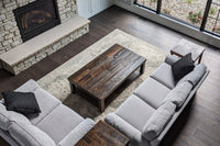 A modern living room with two light-gray sofas faces a handcrafted, customizable reclaimed wood coffee table, set on a large rug beside a stone fireplace and expansive windows. Dark wood floors and minimal decor highlight quality craftsmanship.
