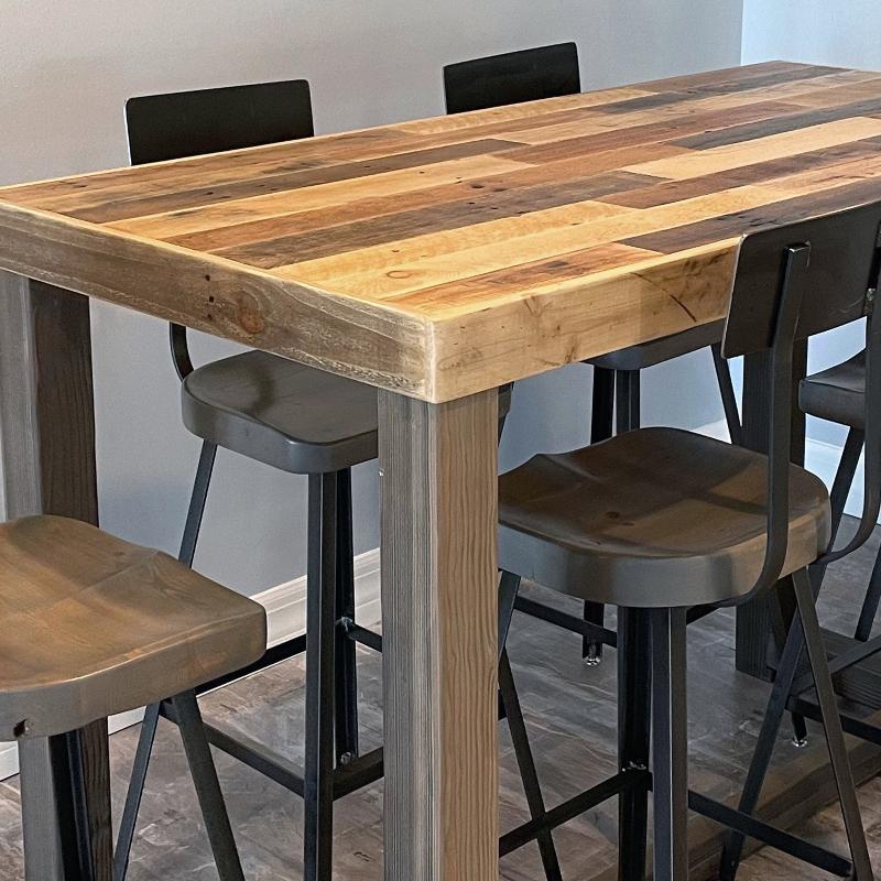 Alt=Bar-height dining table handcrafted from reclaimed wood with a natural, multi-toned finish, paired with four metal bar stools featuring wooden seats and black backs, set on indoor tile flooring.