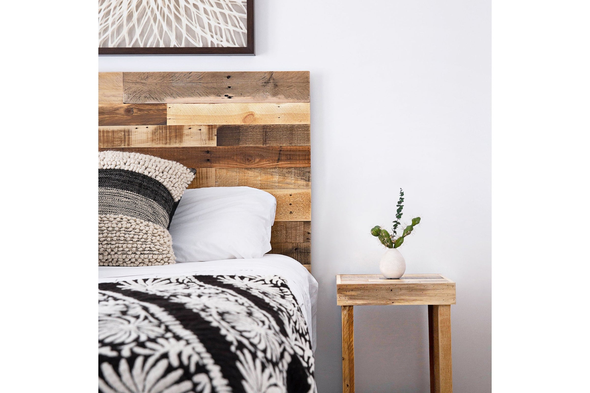 A warm bedroom features a handcrafted, rustic reclaimed wood headboard and textured black and white bedding. A narrow reclaimed wood side table by the bed holds a ceramic vase with greenery, adding timeless quality to the serene space.