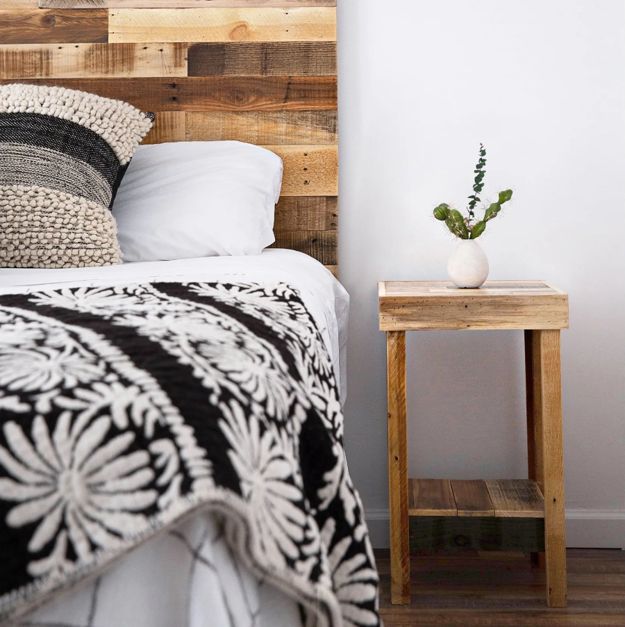 Handcrafted reclaimed wood headboard, white bedding, black and white floral blanket, textured pillow, and sustainable wooden nightstand with vase in a cozy bedroom setting.