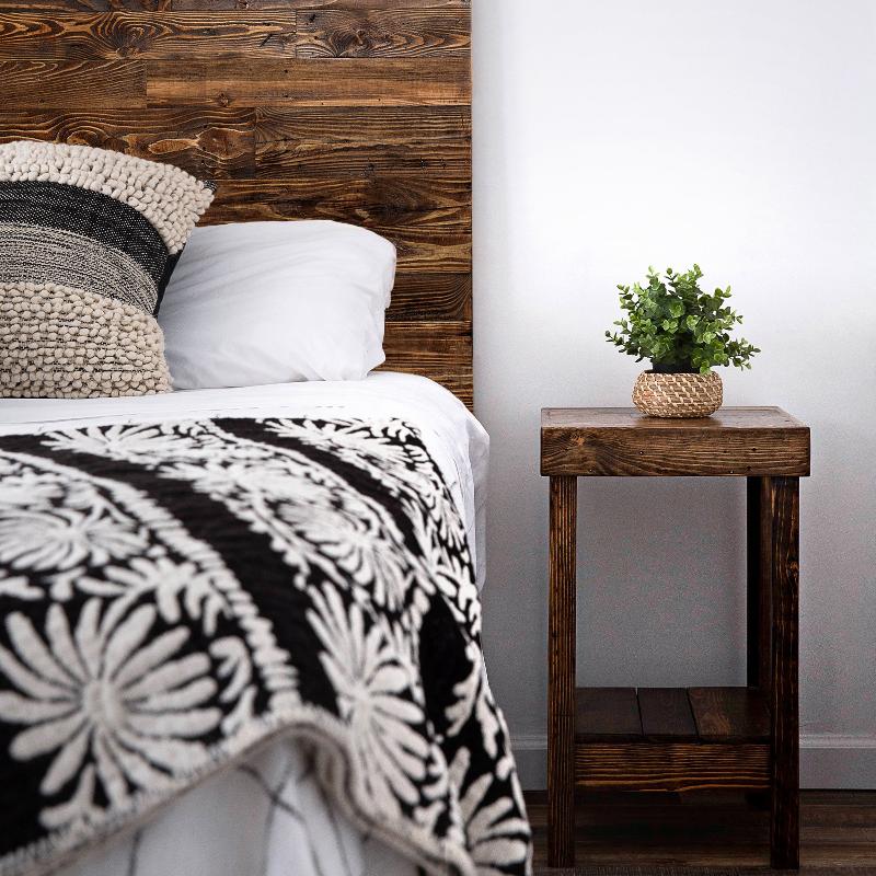 Handcrafted wooden nightstand holds a small potted plant beside a bed with white sheets, textured pillow, and black and white floral blanket in a cozy minimalist bedroom.