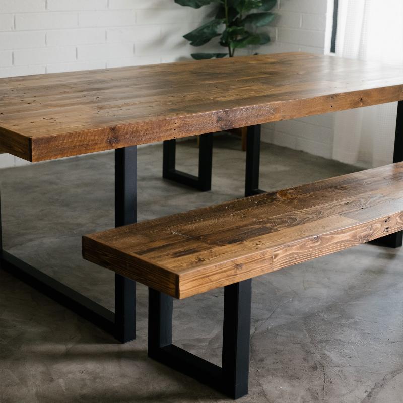 Handcrafted reclaimed wood dining table and matching bench with black steel legs on concrete floor, styled by a white brick wall and potted plant in modern minimalist space. Natural light from window with sheer curtains.