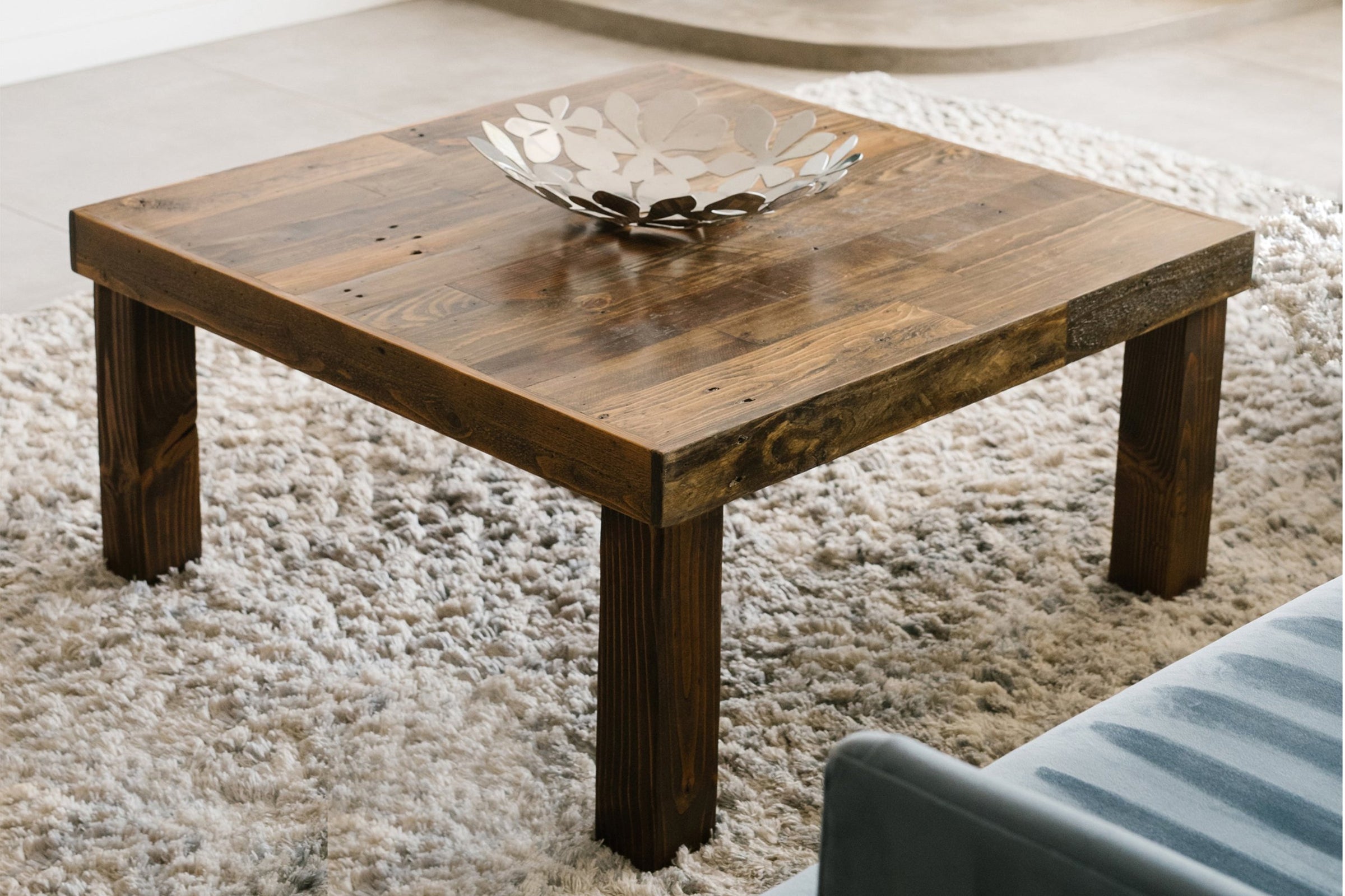 This handcrafted reclaimed wood coffee table, finished naturally for a warm, inviting look, sits atop a plush rug. A metal leaf-pattern bowl accents the centerpiece—perfect as sustainable reclaimed wood furniture.