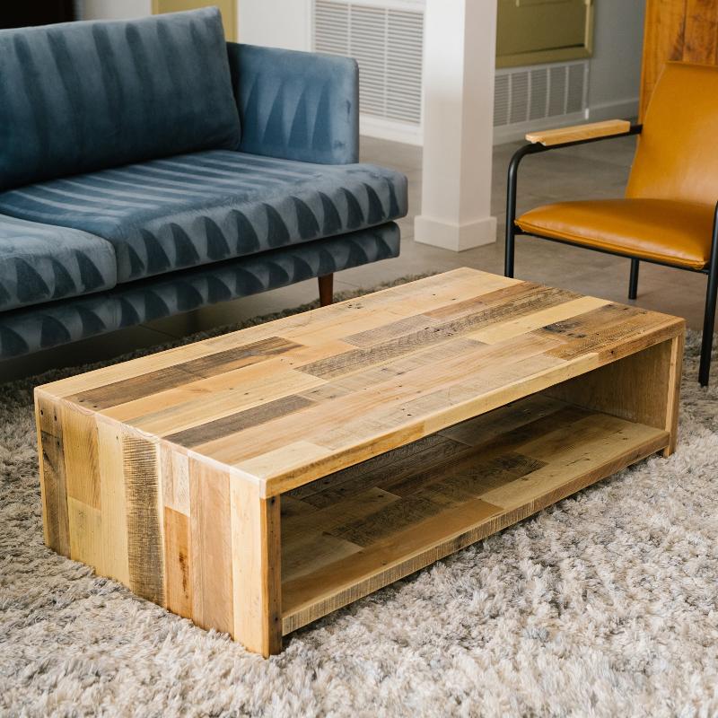 Rectangular reclaimed wood coffee table with open shelf, handcrafted in Arizona, displayed on light textured rug beside blue sofa and tan chair with black steel arms—ideal for modern or historic living rooms.
