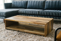 This handcrafted rustic modern coffee table, made from reclaimed wood with an open shelf, brings enduring quality to your living space. Shown on a beige rug before a dark blue sectional, it embodies sustainable furniture design.