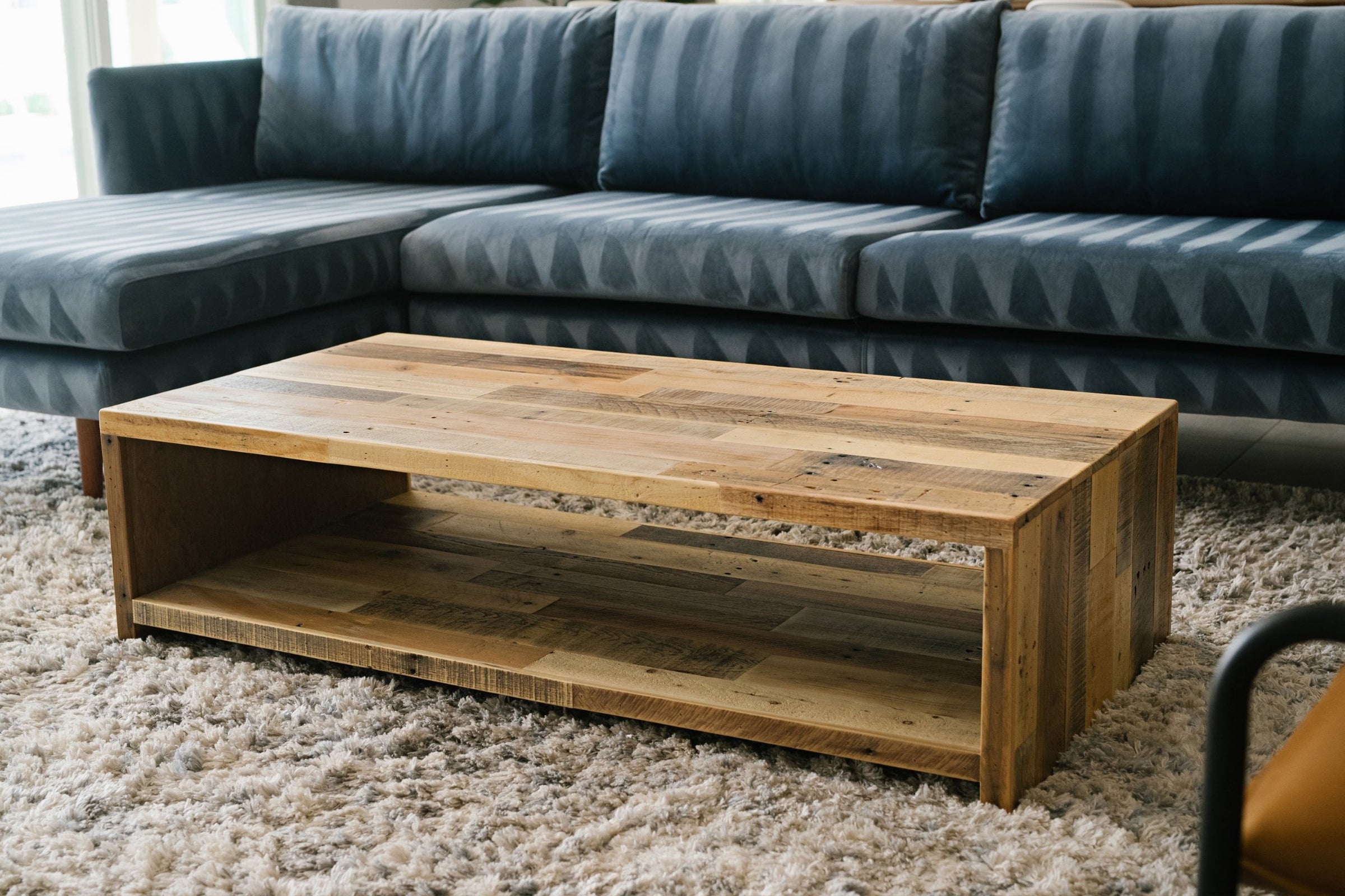 This handcrafted rustic modern coffee table, made from reclaimed wood with an open shelf, brings enduring quality to your living space. Shown on a beige rug before a dark blue sectional, it embodies sustainable furniture design.