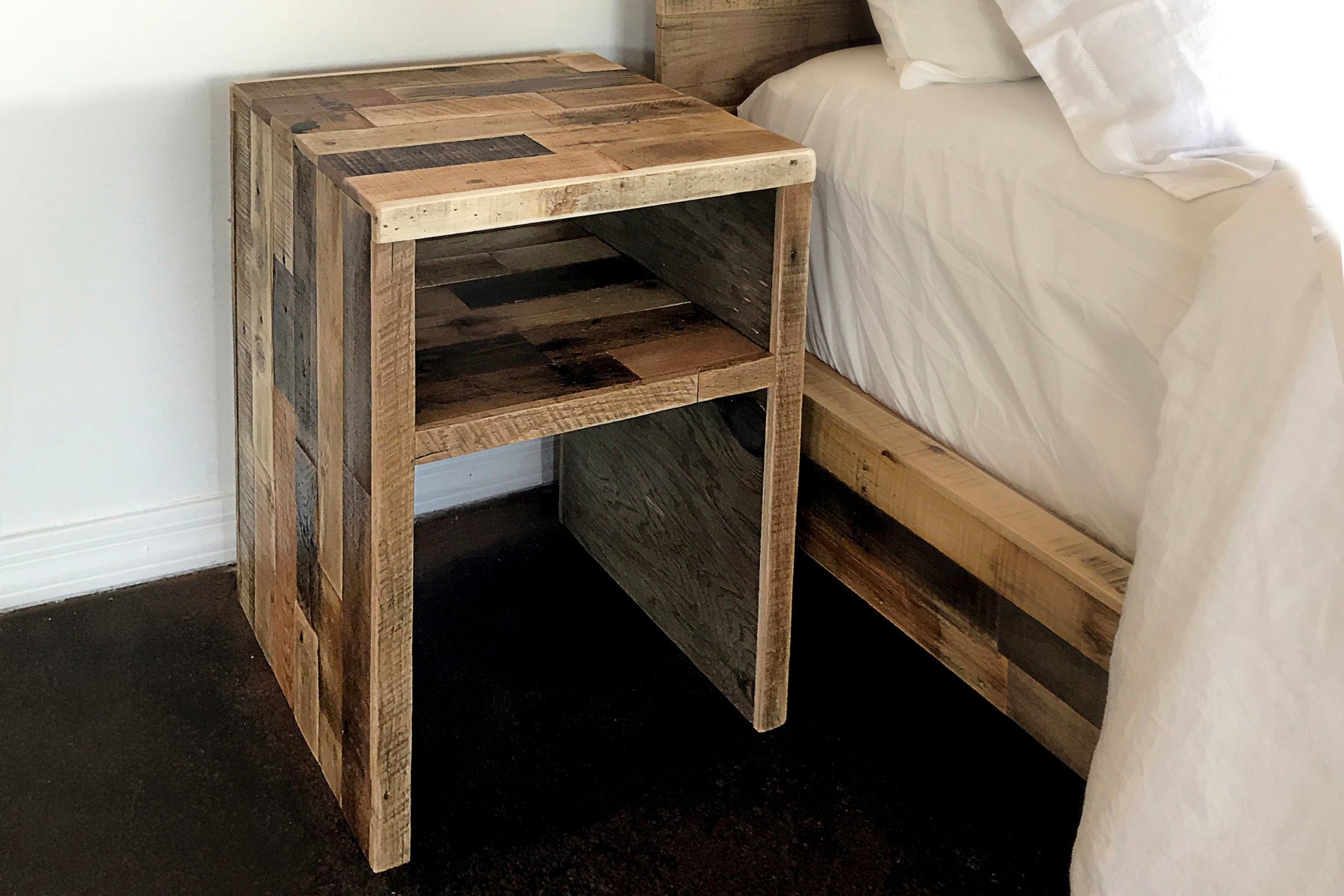 Expertly handcrafted in Arizona, this reclaimed wood bedside table features a patchwork design and open shelf. Perfect as narrow reclaimed wood furniture, it stands beside a bed with white bedding against dark floors and a white wall.