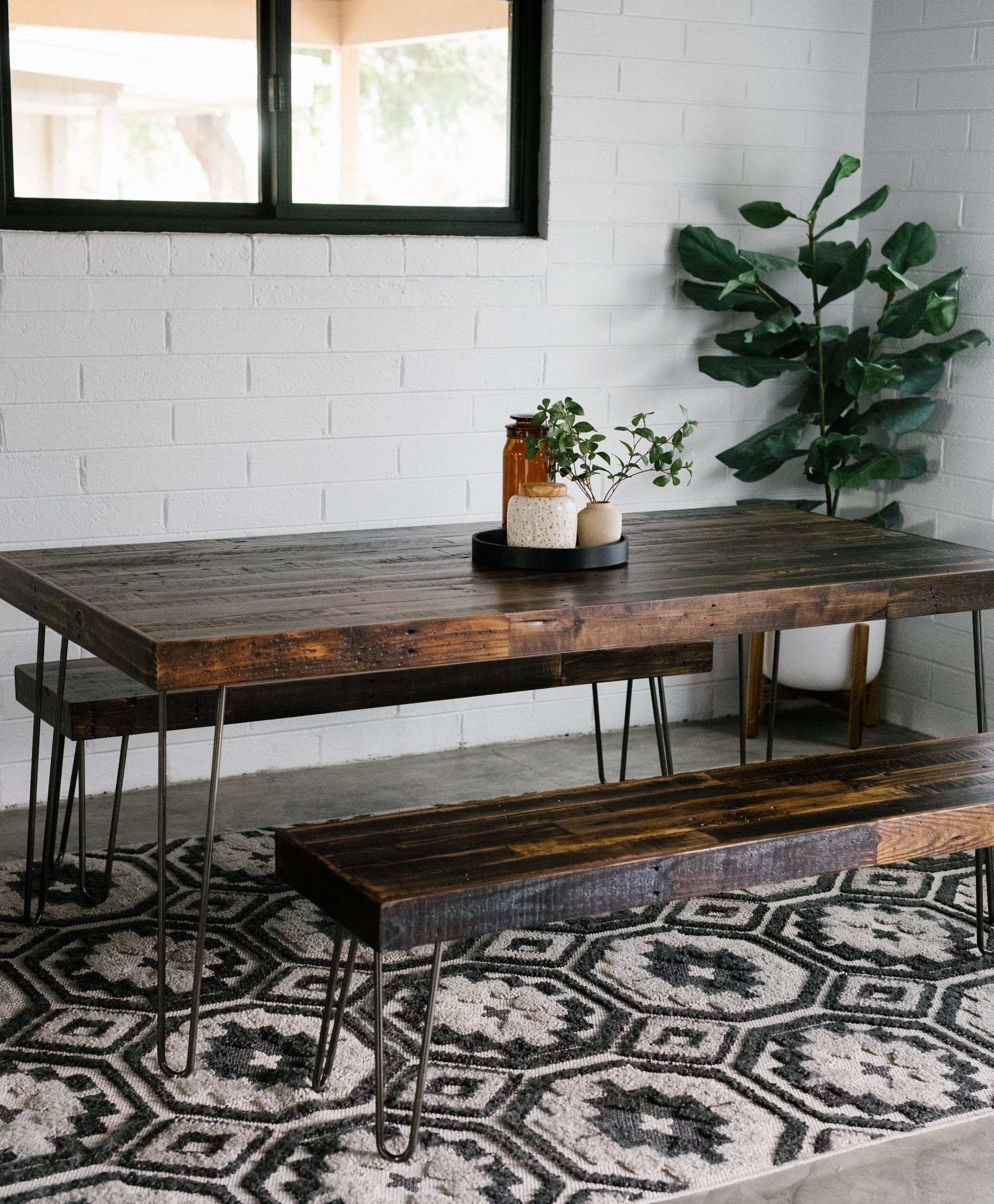 Rustic reclaimed wood dining table with matching benches on patterned rug, potted plants and amber glass jars centerpiece, tall plant in white pot by white brick wall and window; ideal for historic home or modern café.