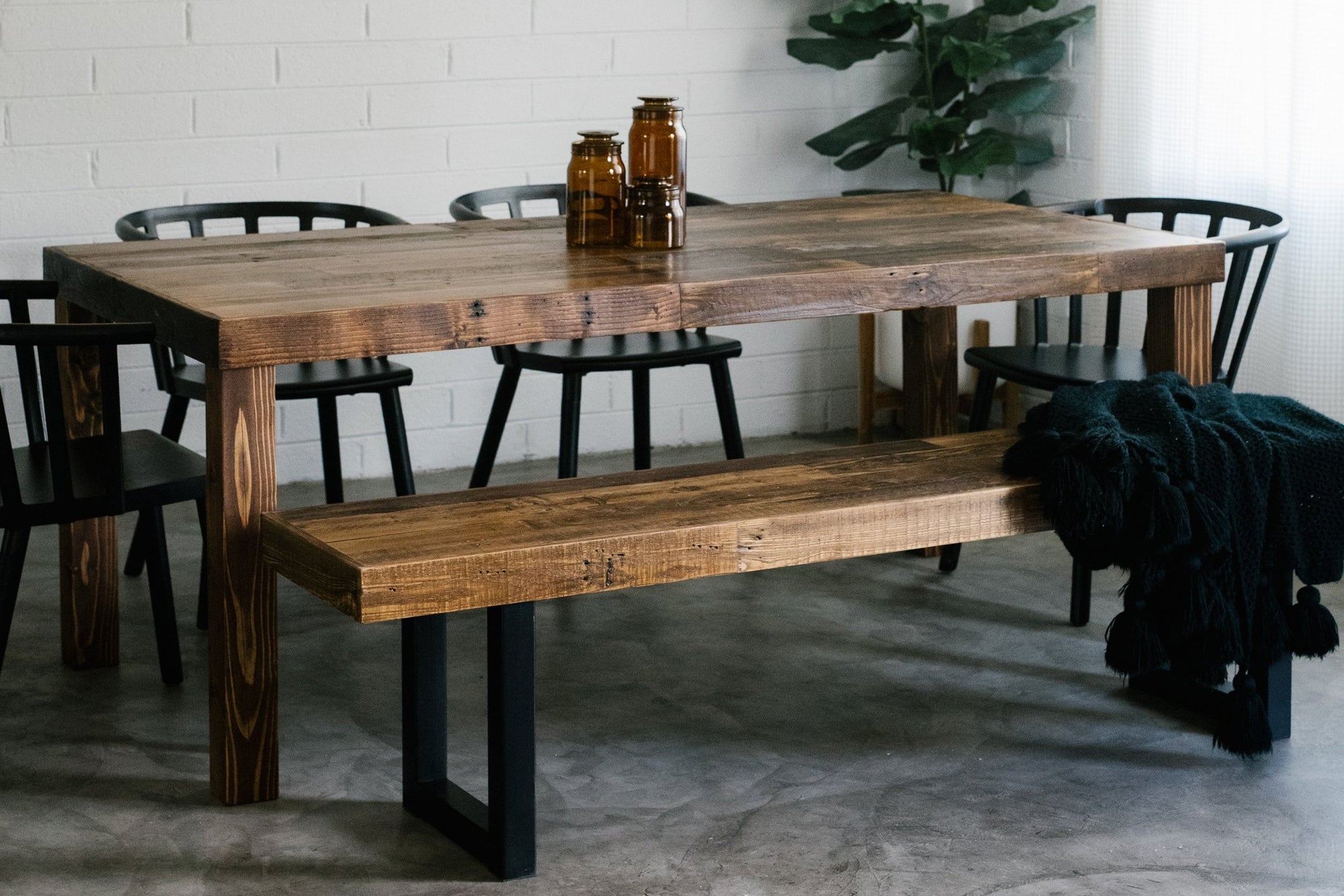 Experience the enduring beauty of a handcrafted reclaimed wood dining table, paired with a matching bench and black chairs atop polished concrete. Amber glass jars and greenery accent this sustainable, artisan-made furniture piece.