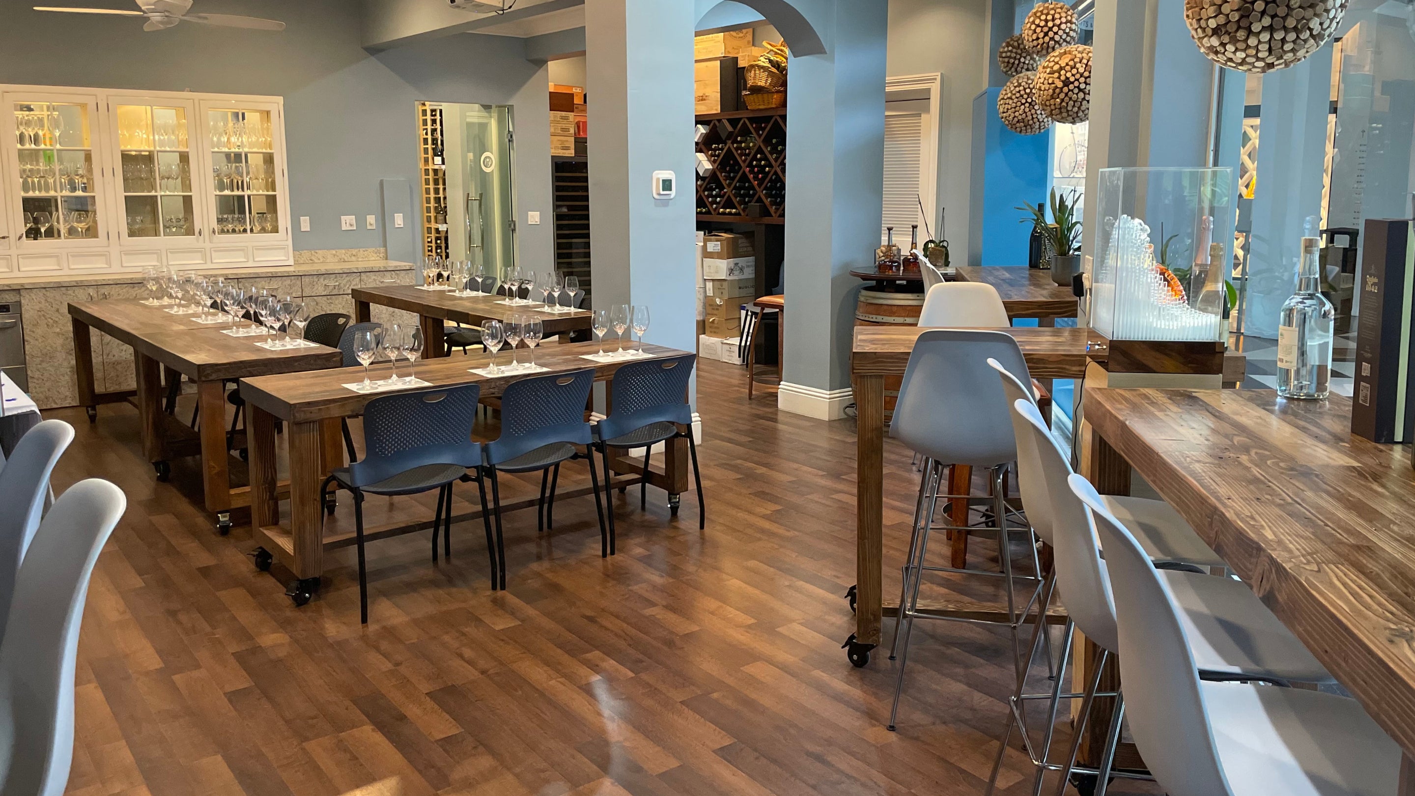 A Florida wine tasting room featuring handcrafted reclaimed wood dining tables and high top bar tables with white barstools, wine racks, blue chairs, and hardwood floors—crafted for lasting quality and timeless style.