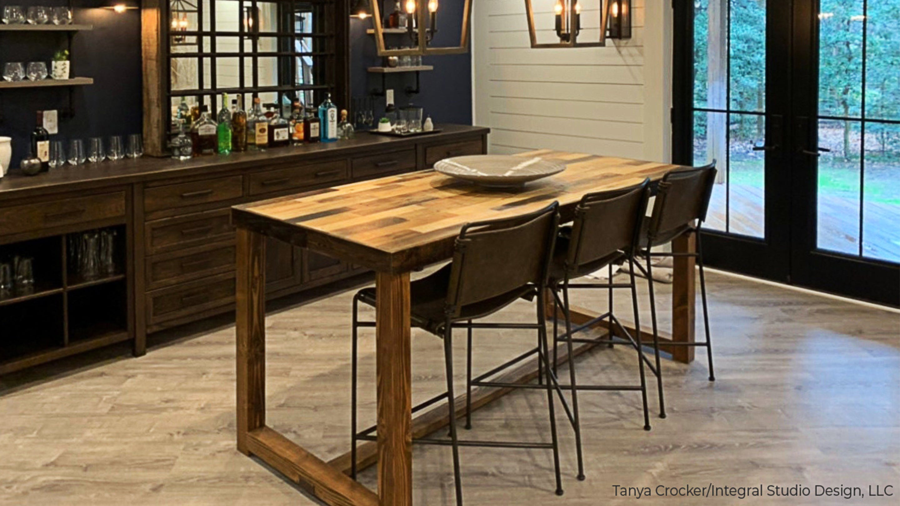 A welcoming home bar area showcases a handcrafted reclaimed solid wood bar table paired with three stools and a dark wood sideboard displaying bottles. Pendant lights and expansive windows highlight enduring craftsmanship.