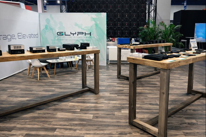 At the trade show, electronic storage devices are showcased on handcrafted reclaimed wood bar-height tables with chairs, complemented by a large GLYPH banner. Warm wooden flooring and potted plants enhance the inviting, quality-focused space.