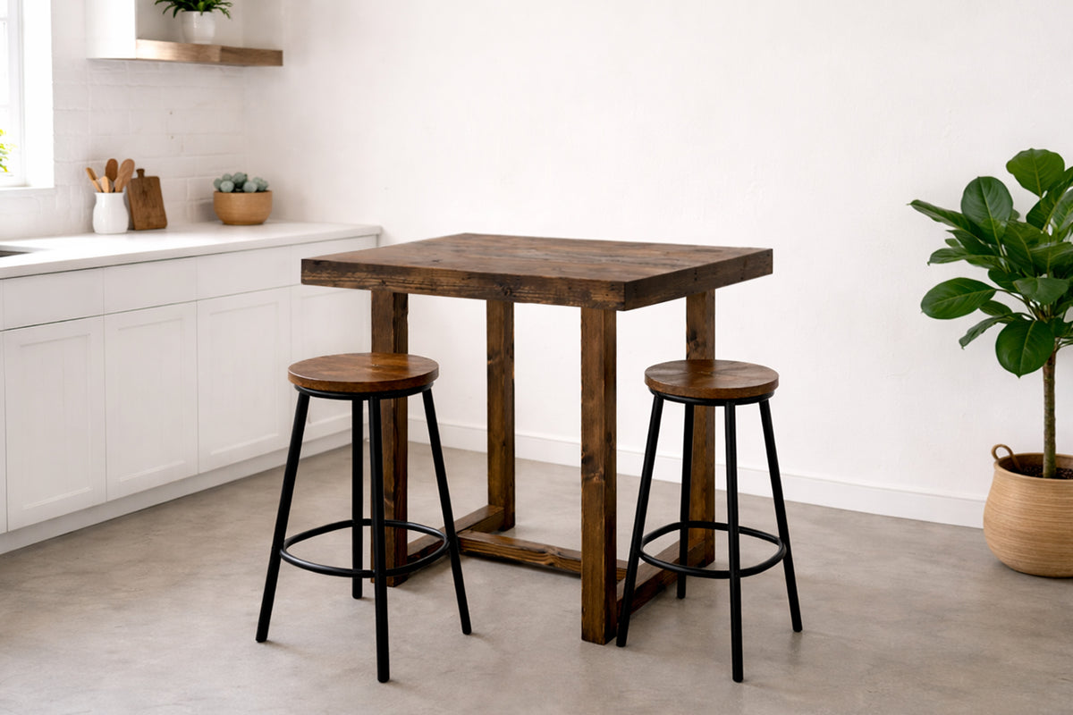 A bright, inviting kitchen features white cabinetry and a handcrafted Arizona reclaimed wood bar-height table. Two wooden bar stools with sturdy black steel legs highlight enduring quality. Natural light and a potted plant enhance the space.
