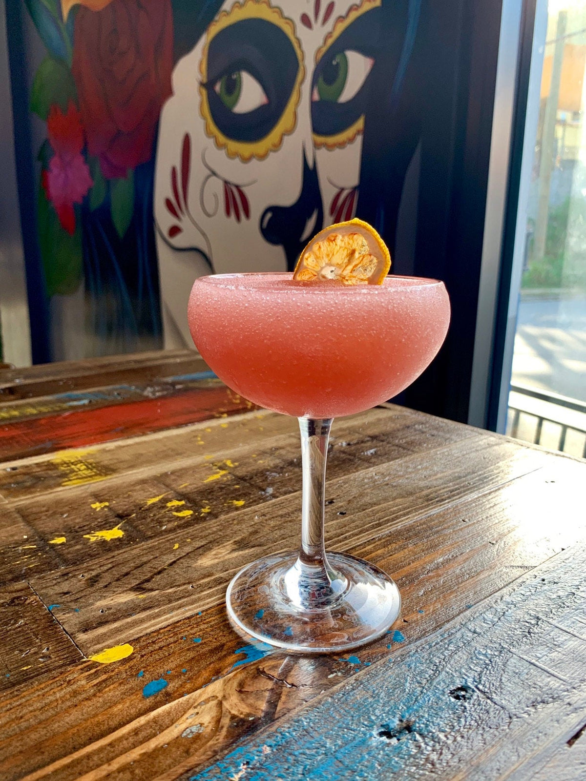 Pink frozen cocktail with dried lime garnish on handcrafted reclaimed wood table; vibrant face and floral mural in backdrop—ideal for modern café or historic home style settings.