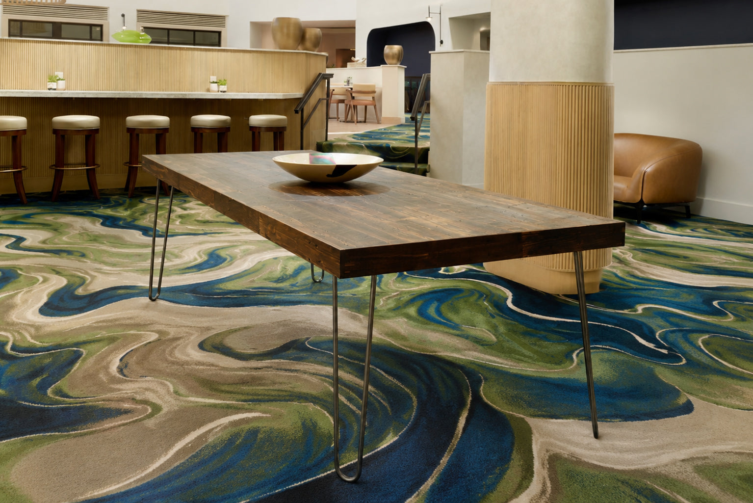 A contemporary space featuring a handcrafted Arizona reclaimed wood bar-height table with sleek hairpin steel legs. Styled with a decorative bowl, bar stools, and artisanal furniture, on a vibrant patterned carpet.