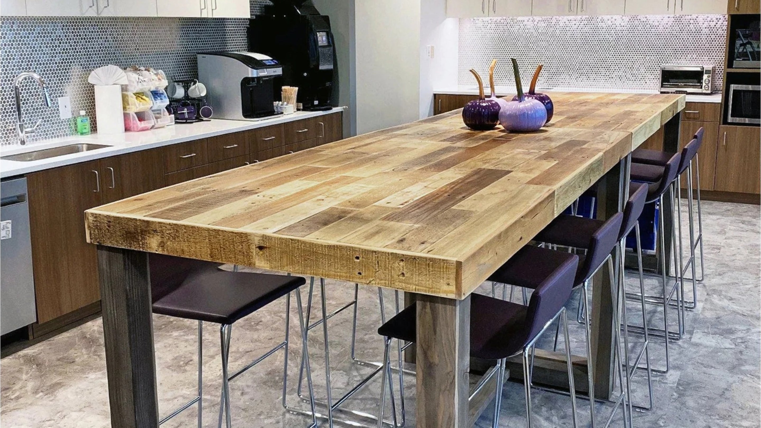 A welcoming office kitchen breakroom features a handcrafted, narrow reclaimed wood bar-height table, paired with sleek purple stools. Light cabinets and a metallic tile backsplash complement this sustainable, Arizona-crafted furniture.