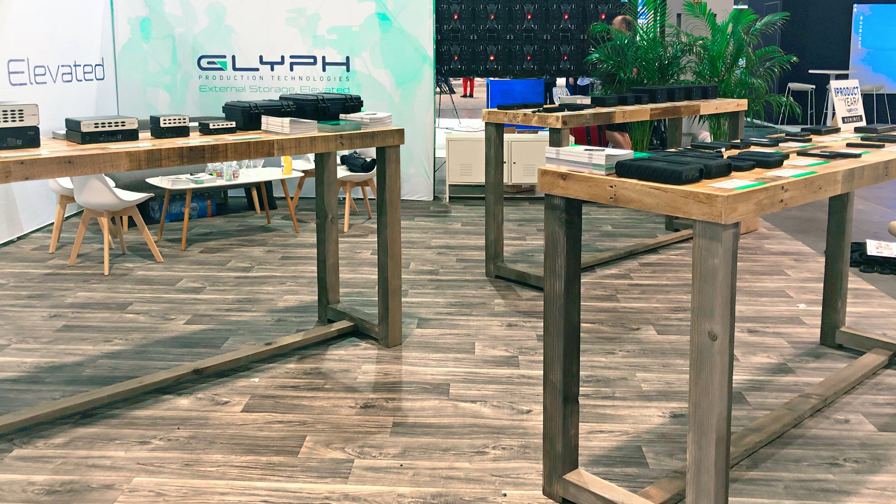 A warm, inviting exhibition booth showcases handcrafted reclaimed wood counter-height tables with a durable steel base, perfect as kitchen islands or restaurant tables. Glyph and Elevated branding enhance the space. Lush plants add a natural touch.