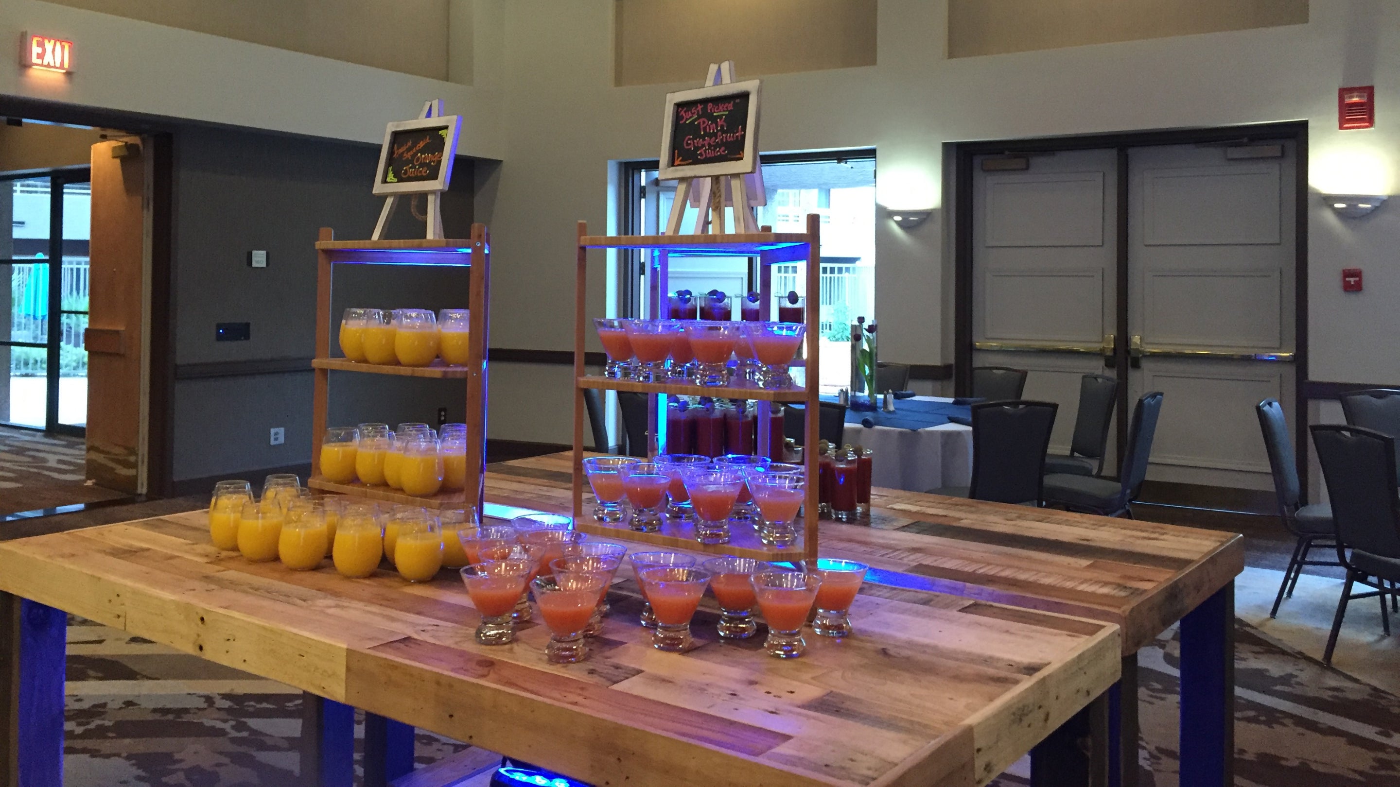 A reclaimed wood counter-height table showcases rows of glass cups with vibrant juices, each labeled by small chalkboard signs. Ideal as a restaurant table or kitchen island, it brings enduring quality to a contemporary venue.