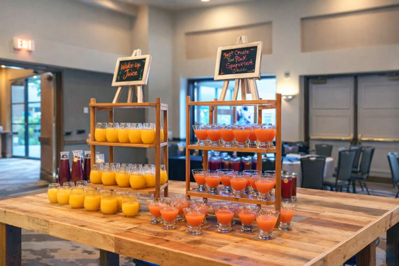 This handcrafted reclaimed wood bar-height table showcases two-tiered wooden racks with rows of orange and red juices. Chalkboard signs highlight Wake-up Juice and Sweet Citrus Pink Grapefruit Splash. Durable, sustainable quality.